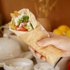 A hand holds a wrapped pita filled with fries, tomatoes, cucumber, and greens, topped with a white sauce. The background features a blurred table setting with dishes and lemons.