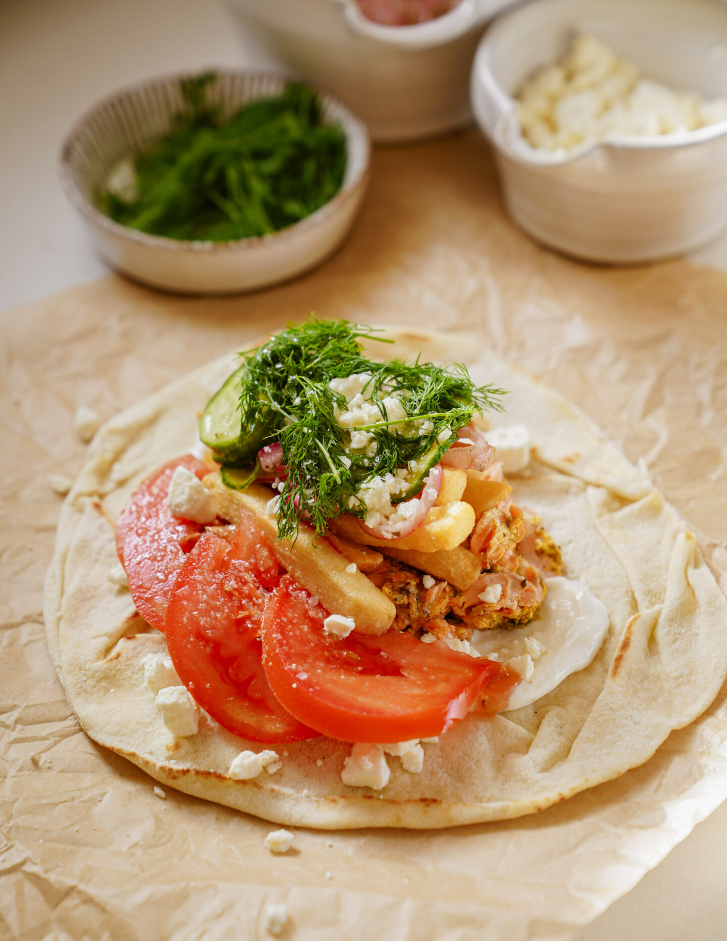 A flatbread topped with sliced tomatoes, pickles, pieces of meat, fries, and crumbled feta cheese. Fresh dill is sprinkled on top. In the background, bowls with herbs and more cheese are visible.