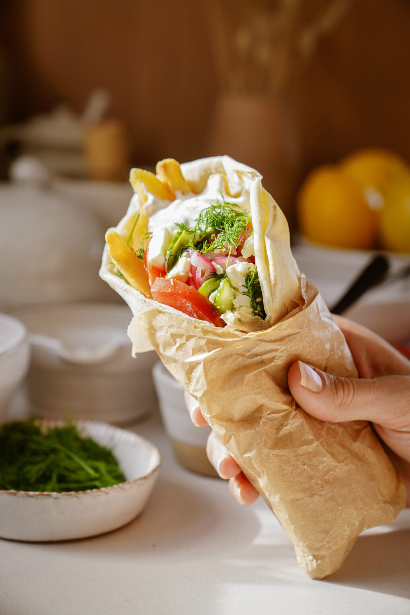 A hand holding a wrapped pita filled with fries, sliced tomatoes, cucumbers, and herbs. A dollop of white sauce is visible on top. In the background, there are plates, bowls, and blurred kitchen items.