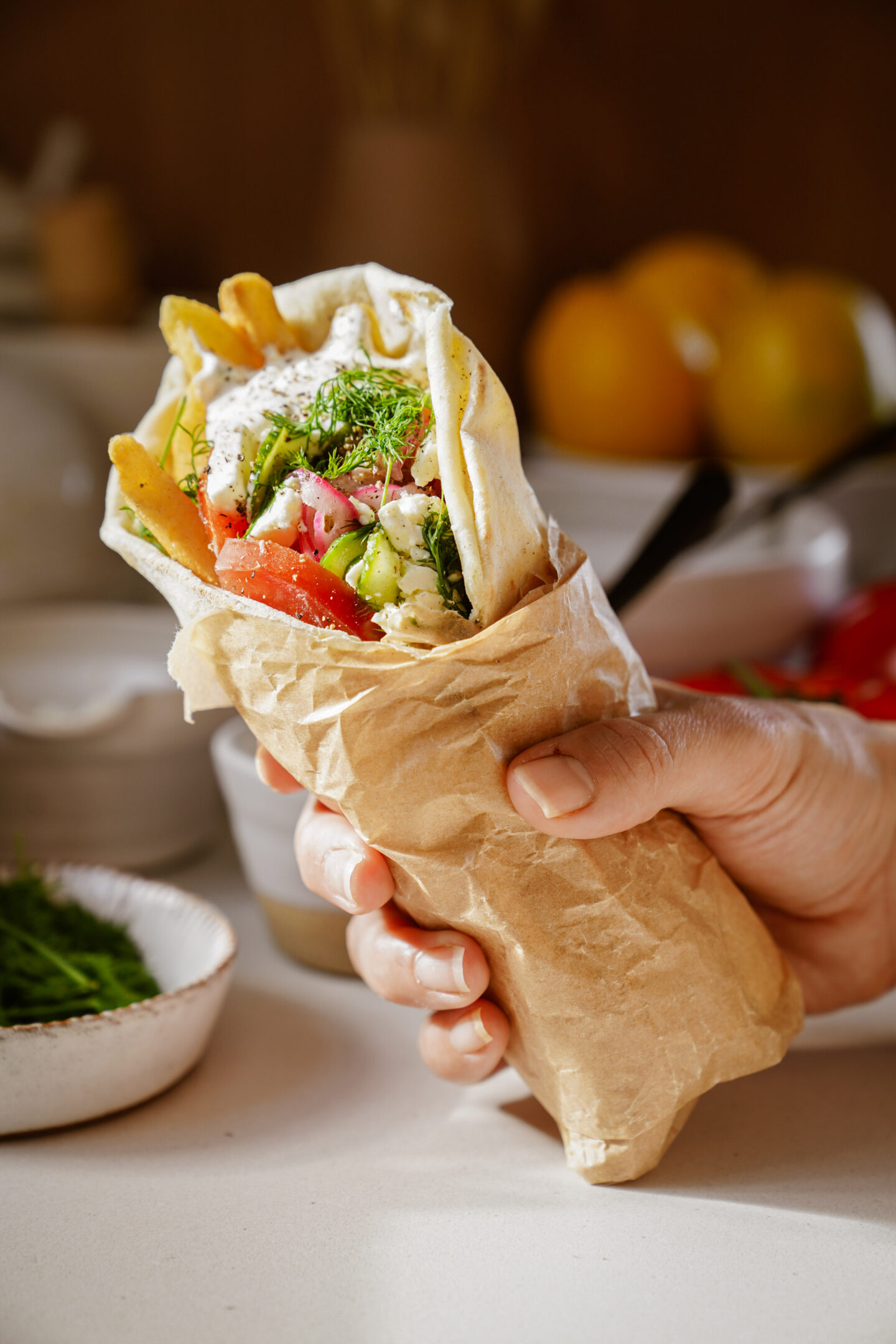 A hand holds a wrapped falafel sandwich filled with ingredients like tomatoes, cucumber, and lettuce, topped with a white sauce and garnished with herbs. The background shows a blurred table with bowls of additional ingredients.