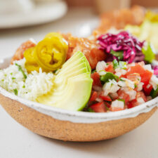 A colorful bowl of food featuring sliced avocado, pickled jalapeños, white rice, diced pico de gallo, shredded red cabbage, and pieces of breaded chicken, all arranged in a rustic ceramic bowl.