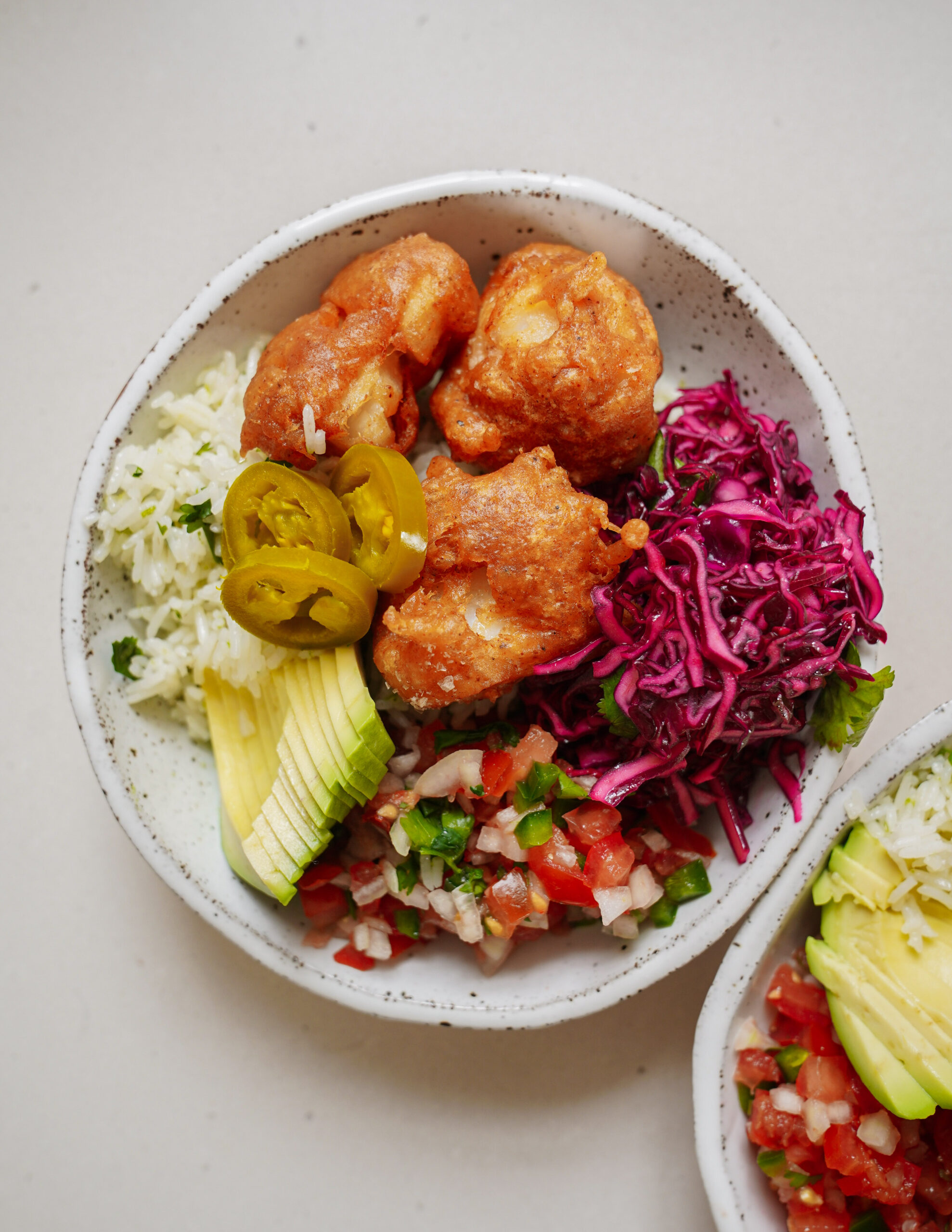 A plate featuring golden fried cauliflower, sliced avocado, pickled jalapeños, shredded purple cabbage, cilantro-lime rice, and fresh salsa. All components are arranged in a white, speckled bowl on a light surface.