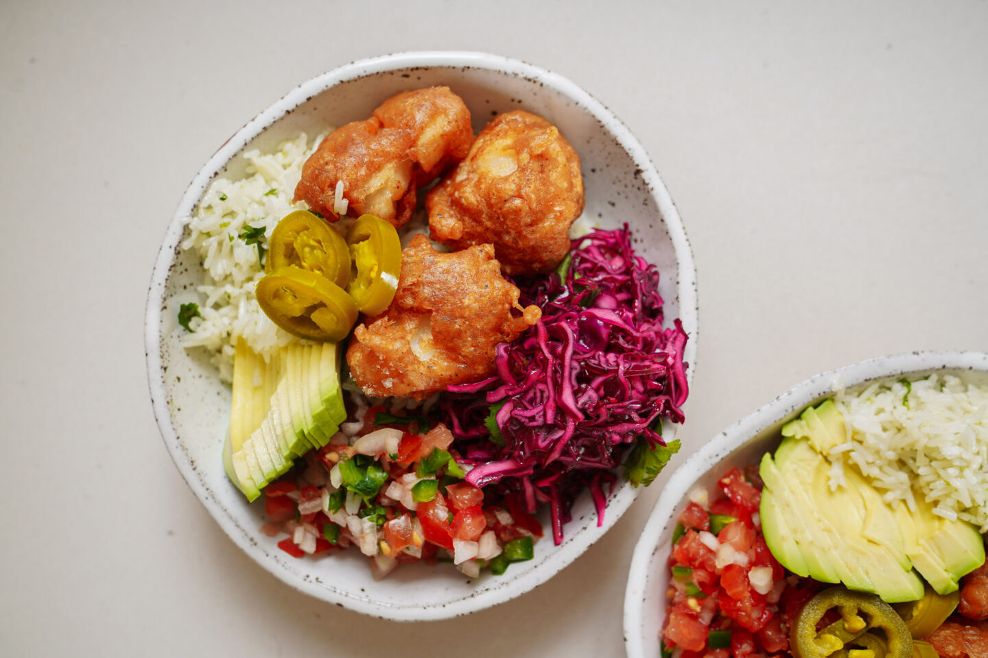 Two white bowls filled with colorful ingredients: crispy fried cauliflower, sliced avocado, jalapeño slices, chopped tomatoes, rice, red cabbage slaw, and fresh greens on a neutral background.