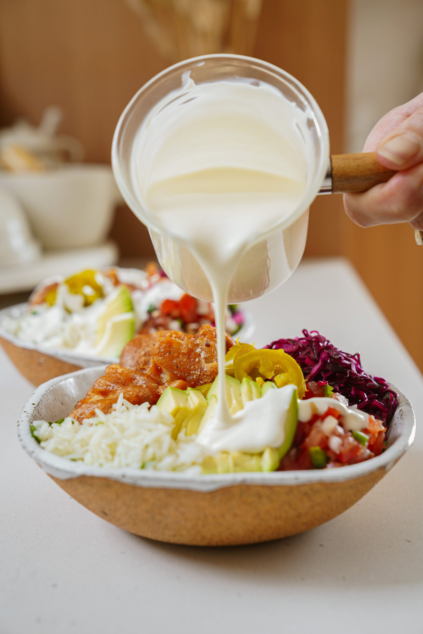 A hand pours a creamy white sauce from a small pan over a bowl filled with rice, diced vegetables, sliced jalapeños, shredded cabbage, and marinated chunks of meat. Another similar bowl is partially visible in the background.