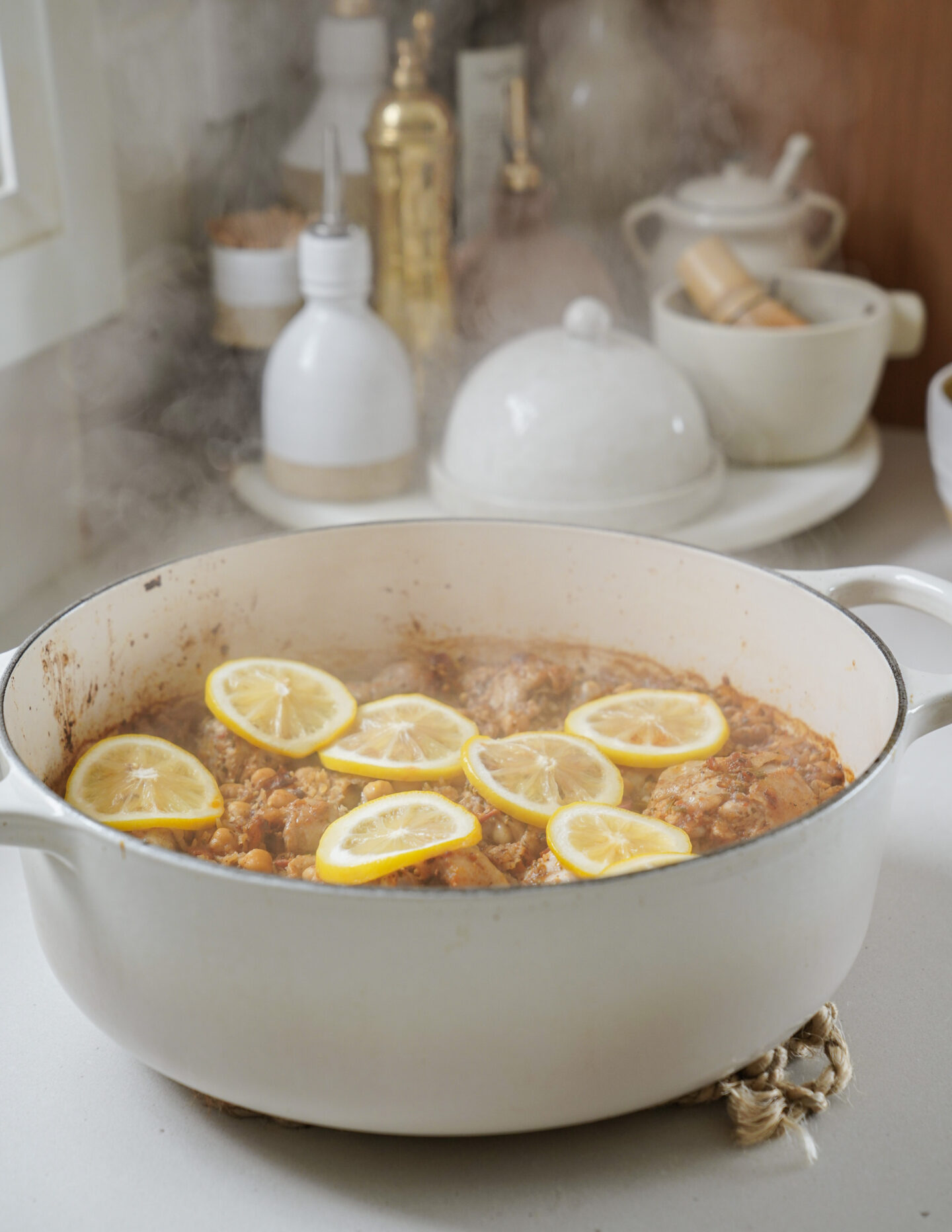 A white pot on a stove contains a steaming dish of chicken and chickpeas, topped with lemon slices. The kitchen counter holds condiments, a mortar and pestle, and a covered dish in the background.