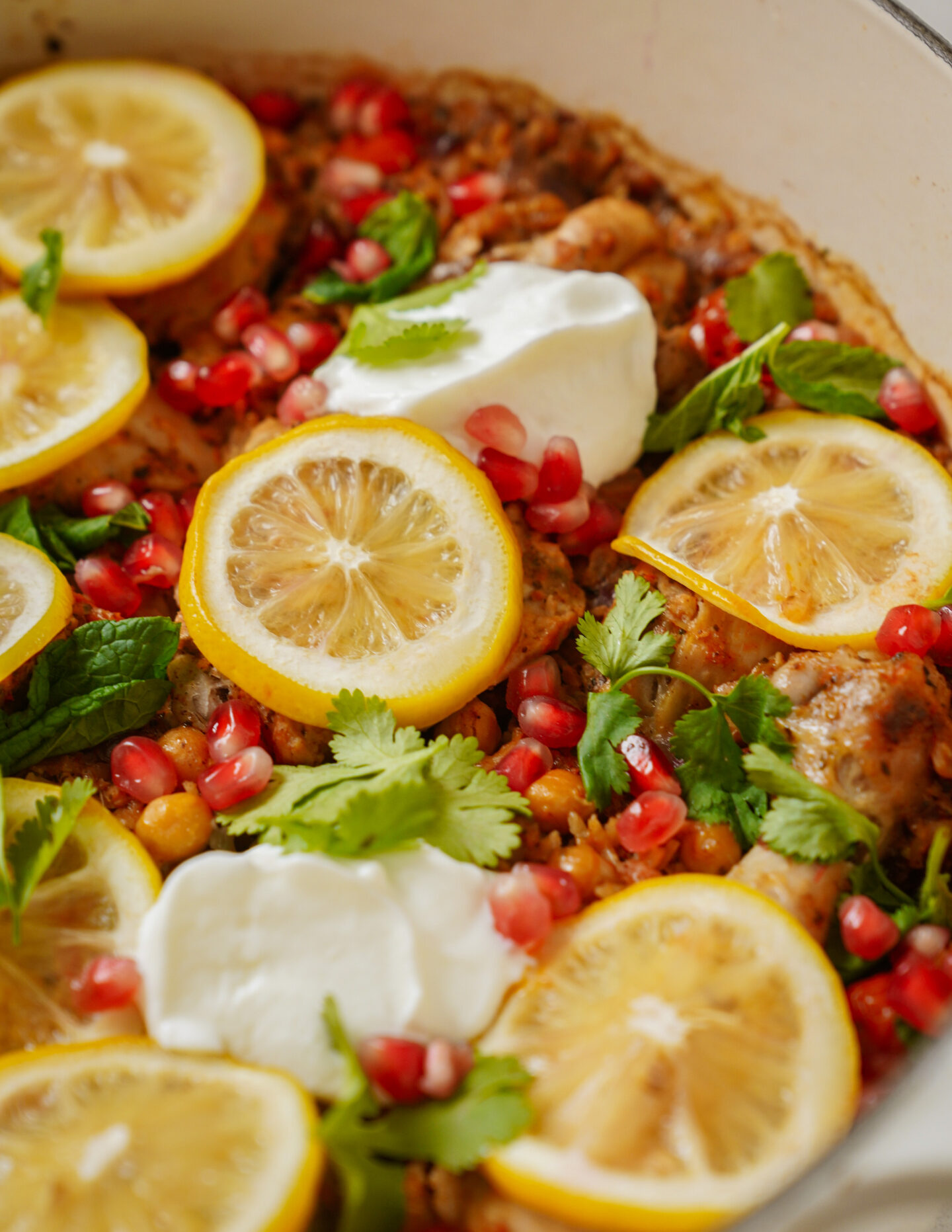 A dish featuring slices of lemon, dollops of yogurt, and a sprinkle of pomegranate seeds on a bed of cooked vegetables and herbs. Fresh mint and cilantro leaves are scattered on top, adding color and texture.