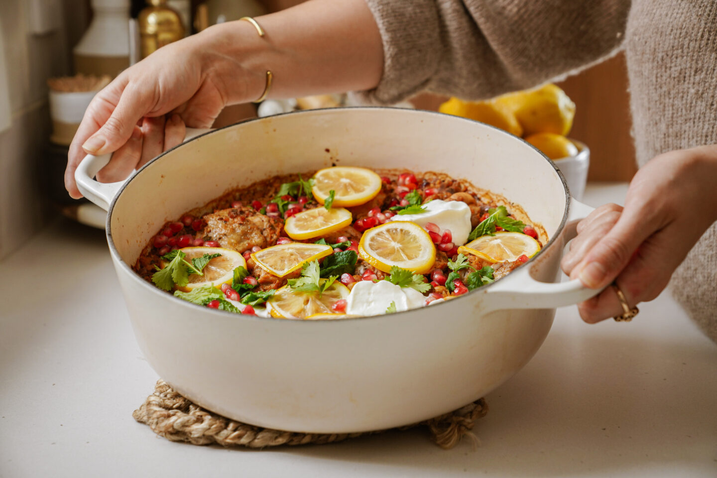 A person holding a white pot containing a baked dish garnished with lemon slices, pomegranate seeds, and greens. The dish is set on a trivet on a countertop, with kitchen items and lemons in the background.