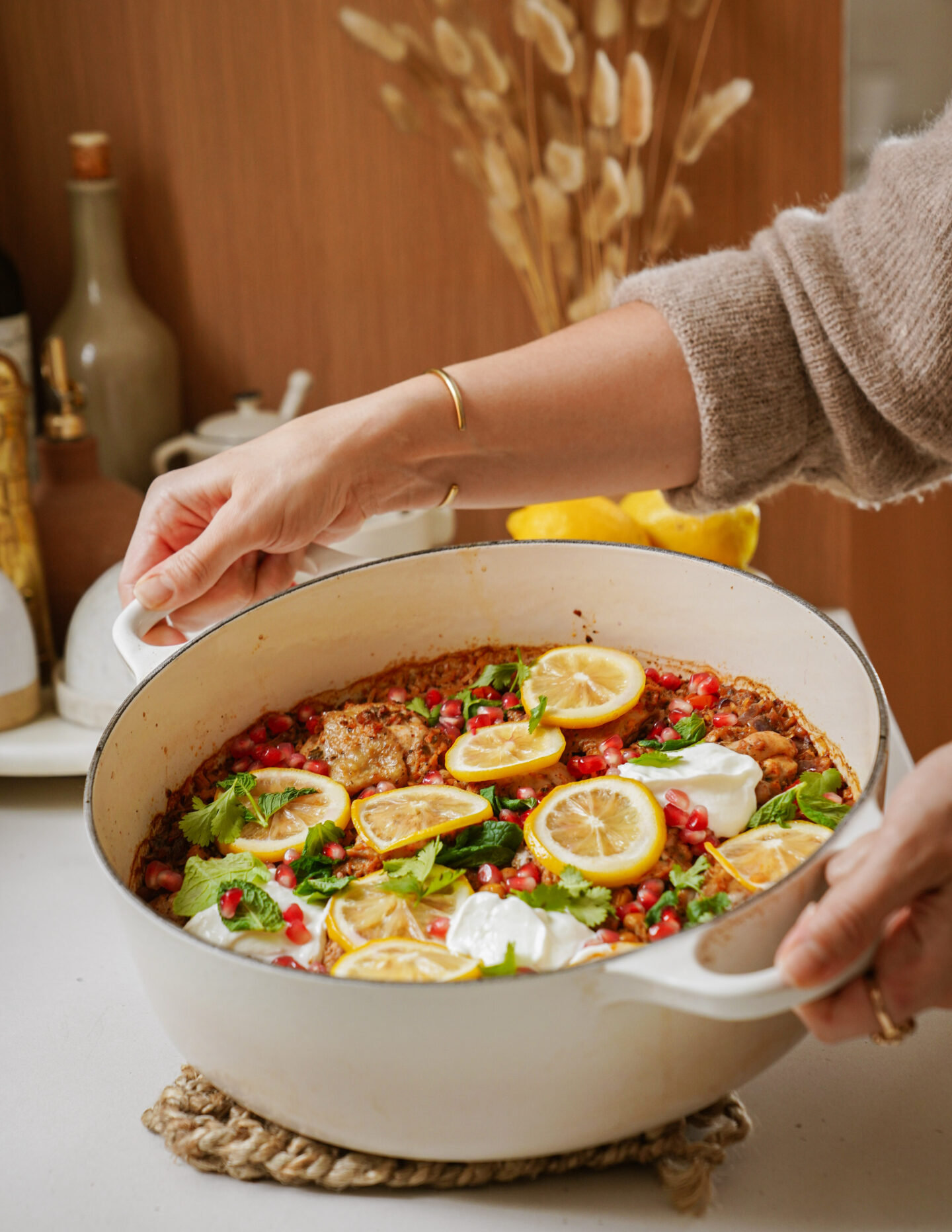 A person is holding a white pot filled with a colorful dish featuring lemon slices, herbs, pomegranate seeds, and dollops of cream. The pot is on a white surface with a neutral-colored kitchen in the background.