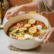 A person holding a white pot filled with a baked dish, topped with lemon slices, dollops of yogurt, pomegranate seeds, and garnished with cilantro. The pot is placed on a woven trivet on a countertop.