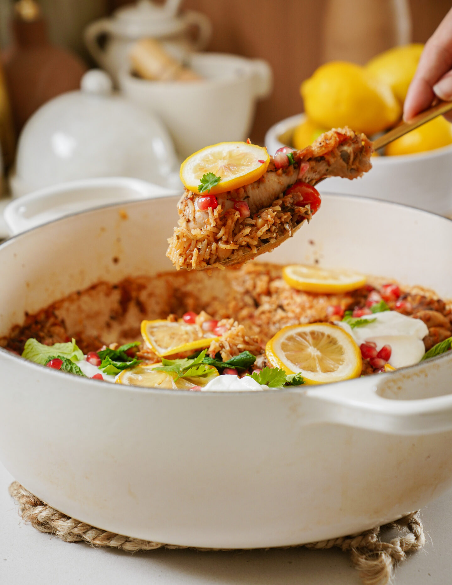 A white pot filled with cooked rice topped with lemon slices, herbs, and pomegranate seeds. A hand holds a spoon with a serving of rice. In the background, lemons and kitchenware are visible.