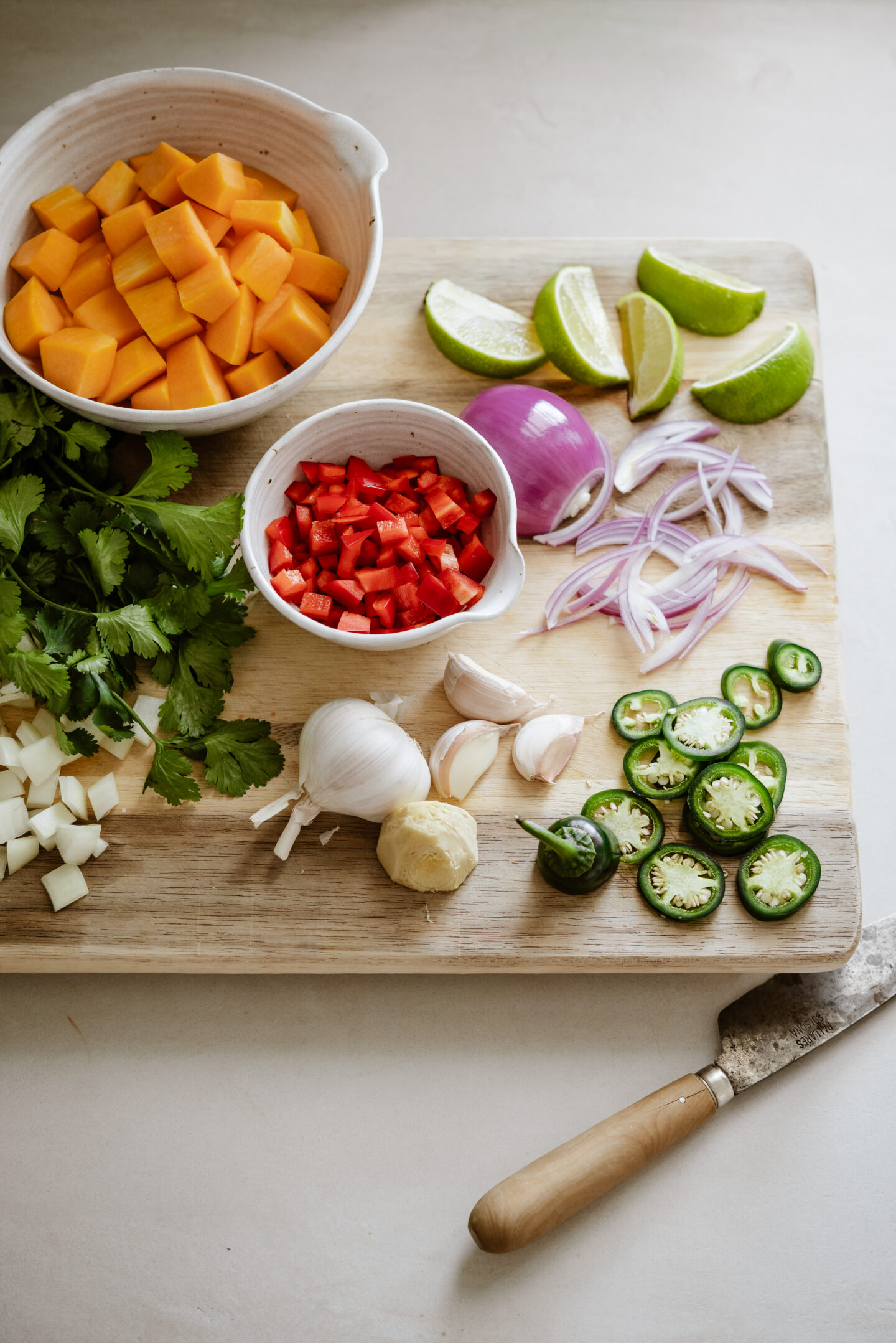 A wooden cutting board with chopped ingredients: cubed squash in a bowl, diced red bell pepper, sliced red onion, halved garlic cloves, sliced jalapeños, lime wedges, cilantro, and peeled ginger. A knife rests beside them.