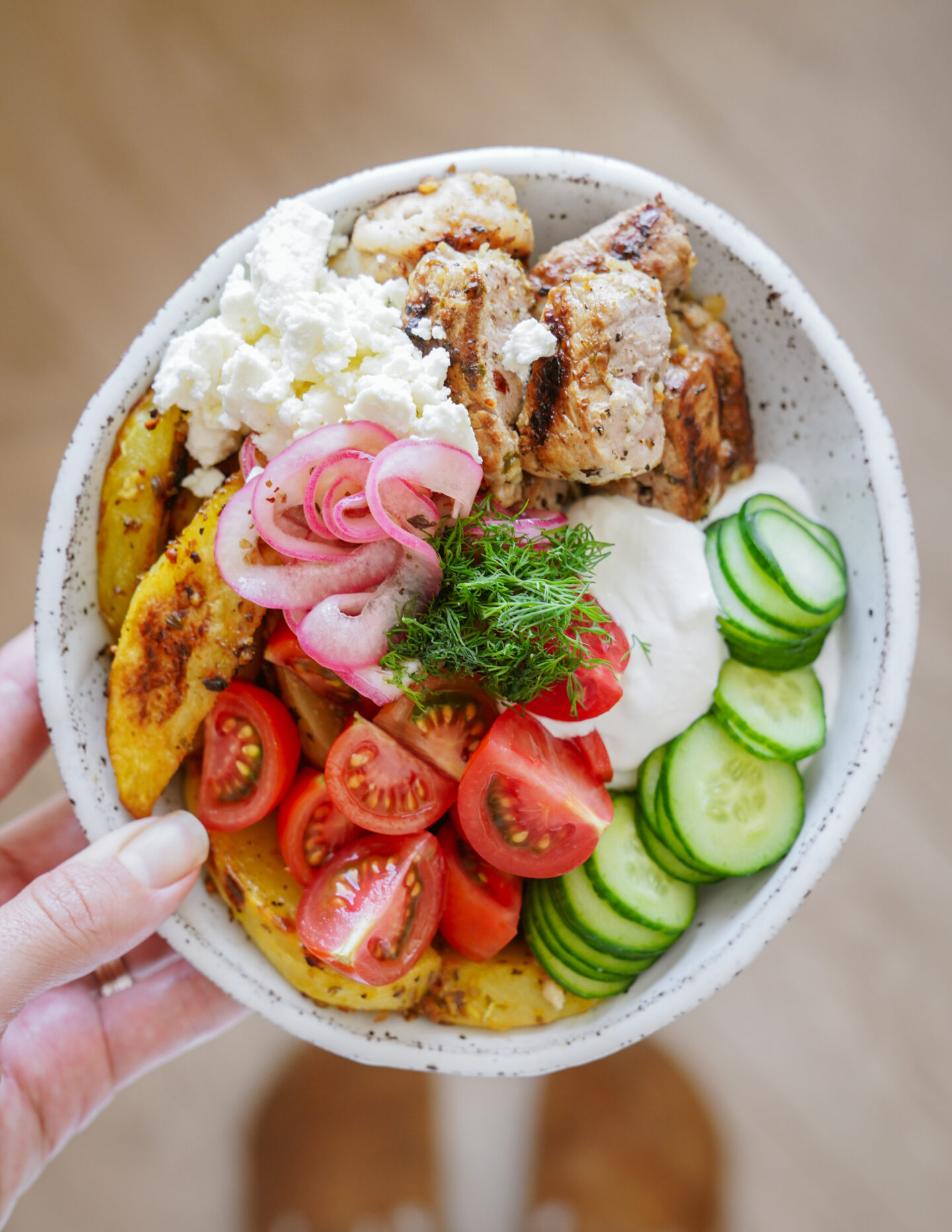 A hand holds a bowl filled with grilled chicken, sliced cherry tomatoes, cucumbers, pickled onions, roasted potato wedges, a dollop of white sauce, and a topping of crumbled feta cheese and fresh dill.