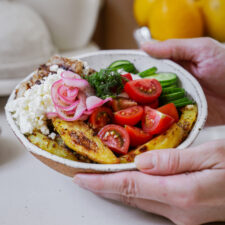 Close-up of hands holding a bowl filled with roasted potato wedges, cherry tomatoes, sliced cucumbers, pickled onions, crumbled feta cheese, and a sprig of dill. A lemon and kitchen items are blurred in the background.