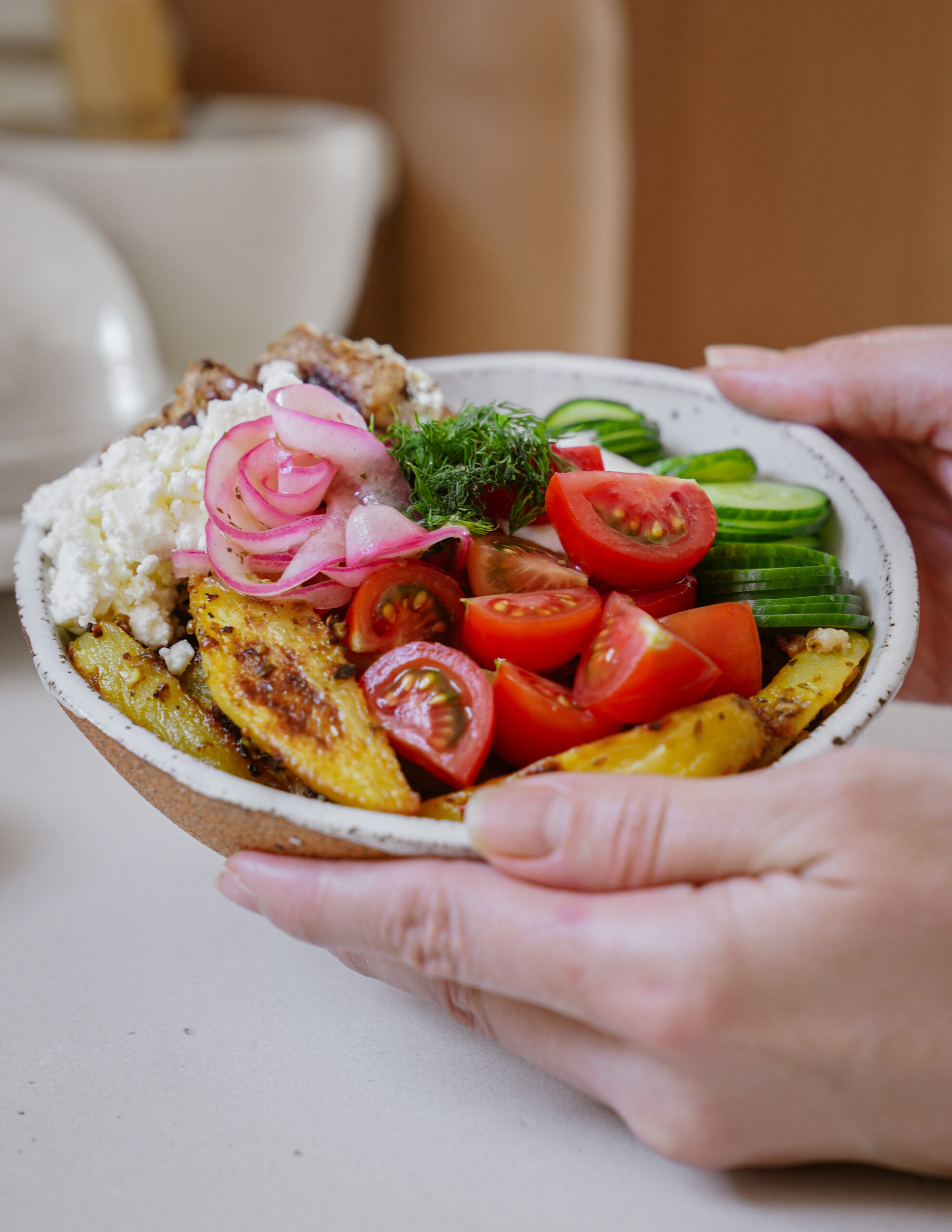 Hands holding a bowl with sliced cucumbers, cherry tomatoes, red onion, cottage cheese, dill, and seasoned potato wedges.