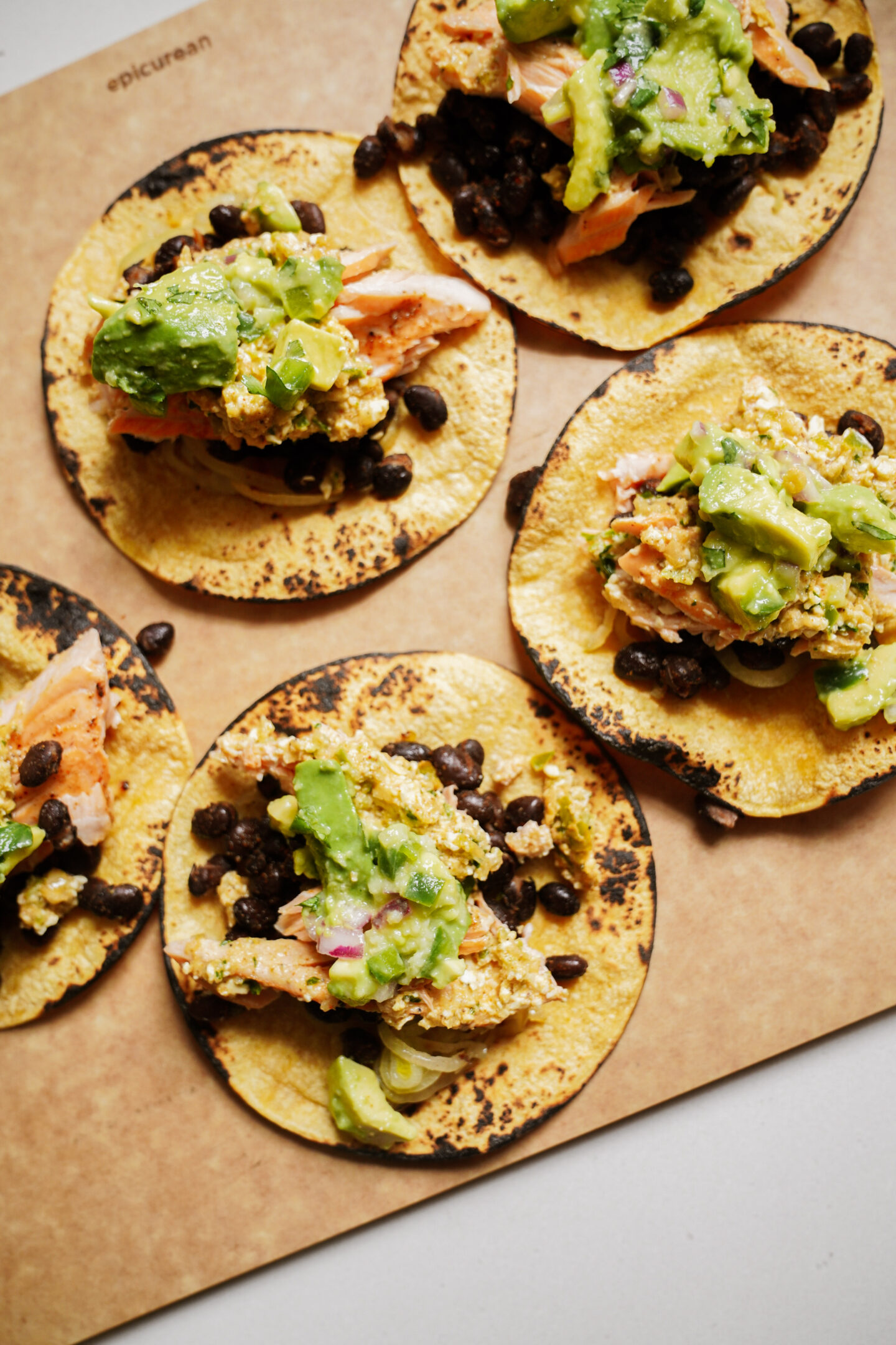 Five corn tortillas with black beans, scrambled eggs, and avocado on a wooden board. The tortillas appear lightly charred, and the toppings are evenly distributed.