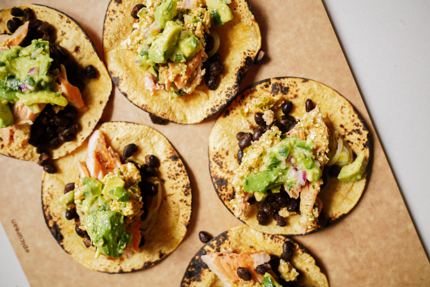 Five corn tortillas with black beans, scrambled eggs, and avocado on a wooden board. The tortillas appear lightly charred, and the toppings are evenly distributed.