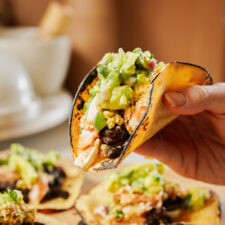 A hand holding a taco with a charred tortilla filled with black beans, chicken, cheese, and guacamole. More tacos on a blurred background, on a light-colored table.