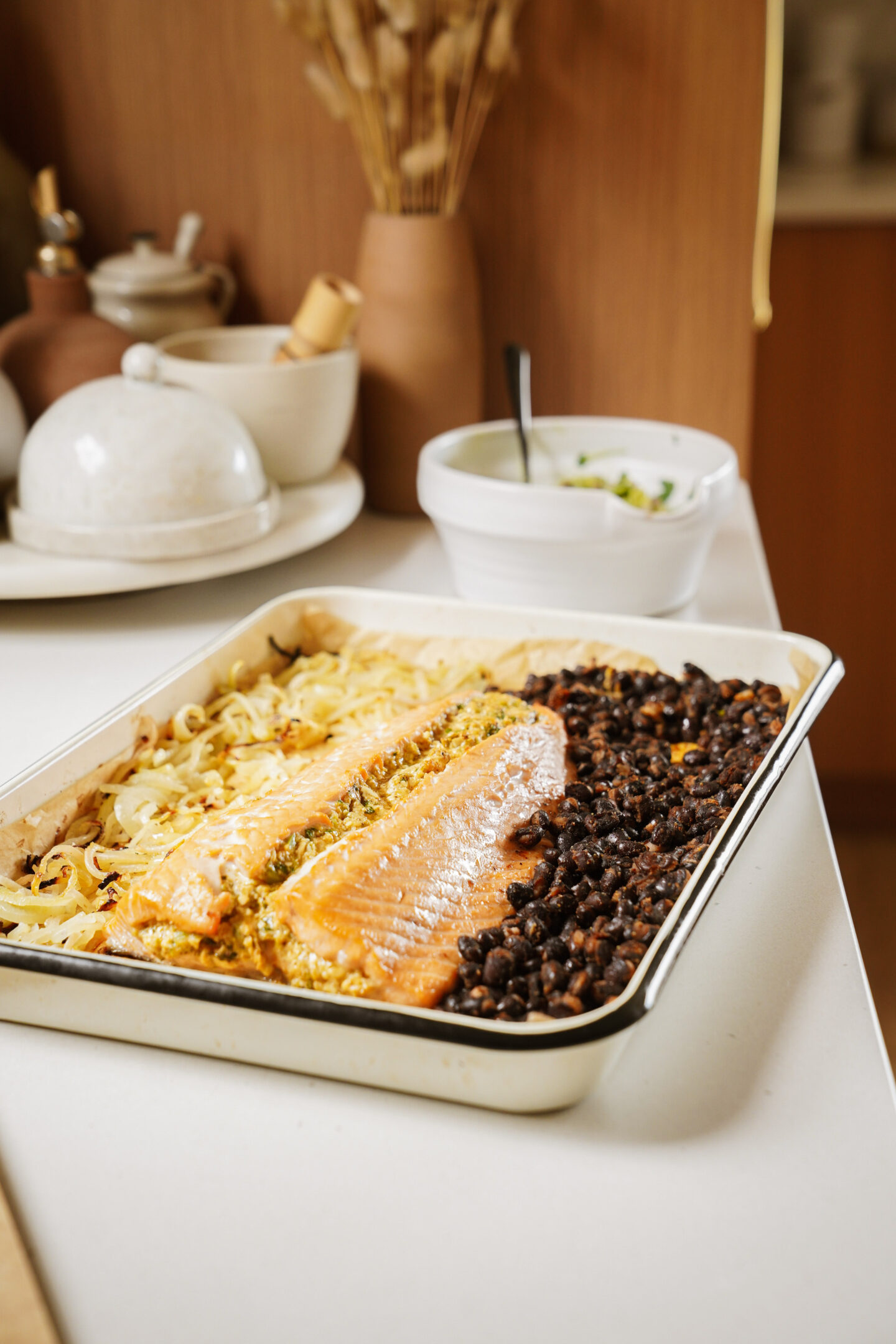 A tray of baked salmon is flanked by sautéed cabbage on one side and black beans on the other. The tray is placed on a kitchen counter with ceramic dishes, a plant, and a white bowl with a green salad in the background.