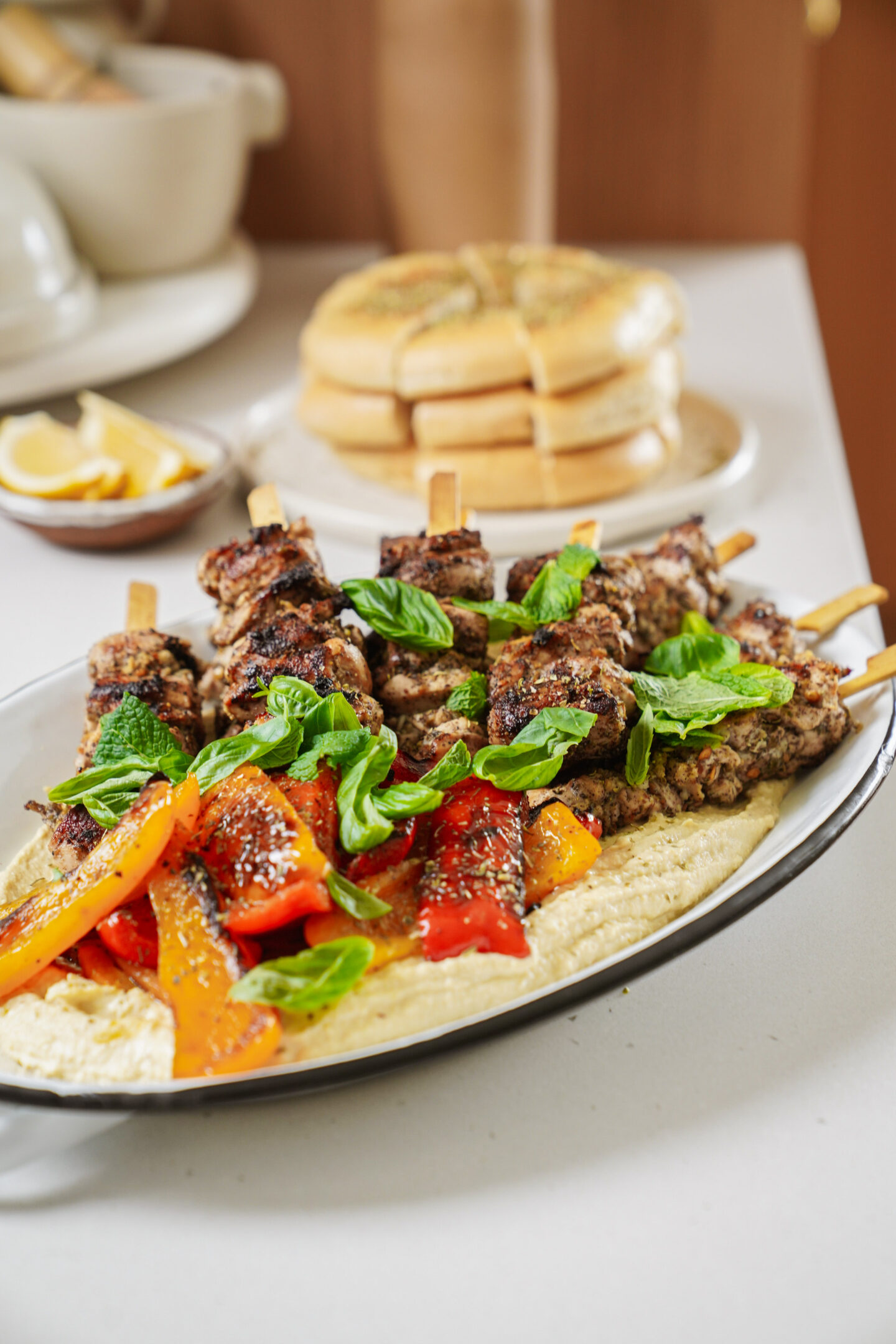 A plate of grilled meat skewers garnished with fresh basil sits atop a bed of hummus, accompanied by grilled red and yellow bell peppers. Lemon wedges are in a small bowl nearby, and a stack of pita bread is in the background.