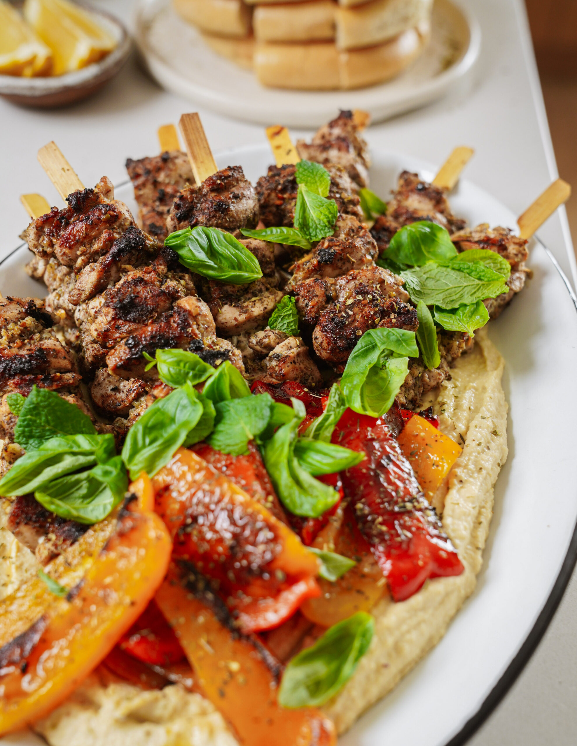 Grilled meat skewers garnished with fresh basil sit atop a bed of hummus. Accompanied by roasted red and yellow bell peppers on a white plate. A stack of pita bread is visible in the background.