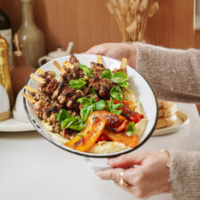 A person holds a plate of grilled meat skewers garnished with fresh basil leaves, served over hummus and roasted red and yellow bell peppers. The background shows various kitchen items.