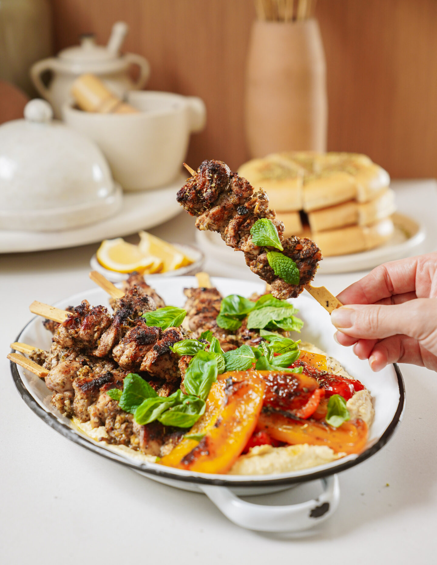 Grilled skewered meat garnished with mint and basil is served on a platter with roasted bell peppers and hummus. A hand holds one skewer. In the background, theres a stack of pita bread and a ceramic teapot set.